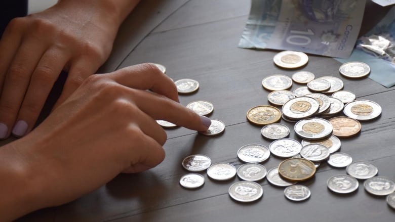 two hands count canadian coins on a wood table with two bills in the background.