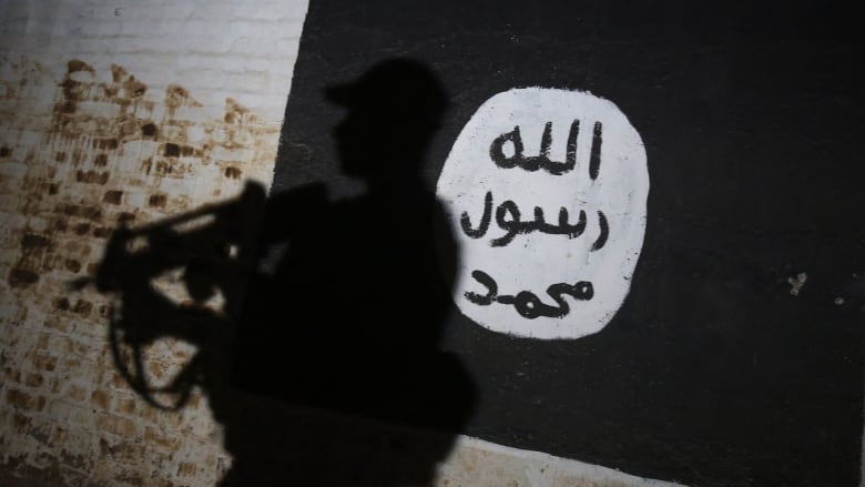 A shadow of a man carrying an assault rifle is cast on a painted black flag with Arabic script.