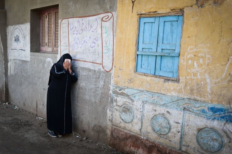 A woman holds her face in her hands and leans against a wall.