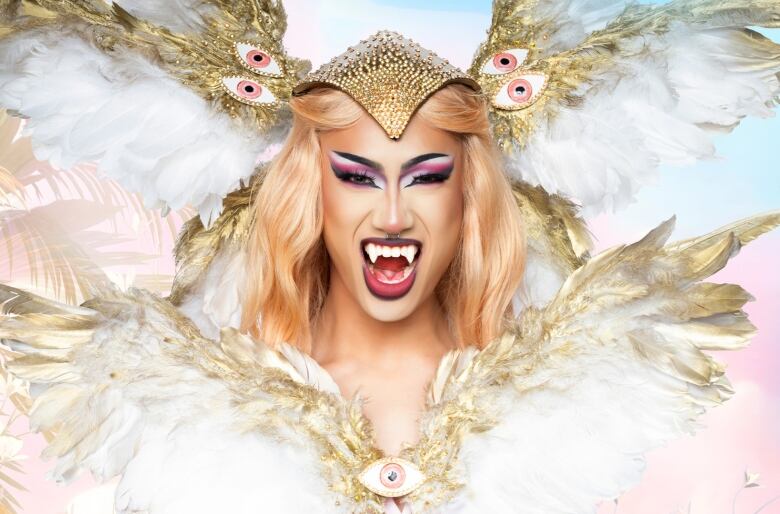 A drag queen in a blond wig and gold helmet with feathers strikes a pose while wearing fangs. 