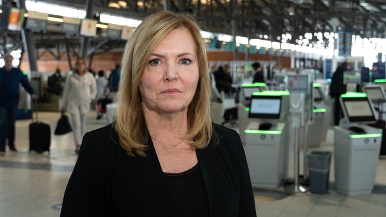 A women in a black blazer with blonde hair stands in front of the check-in machines at an airport