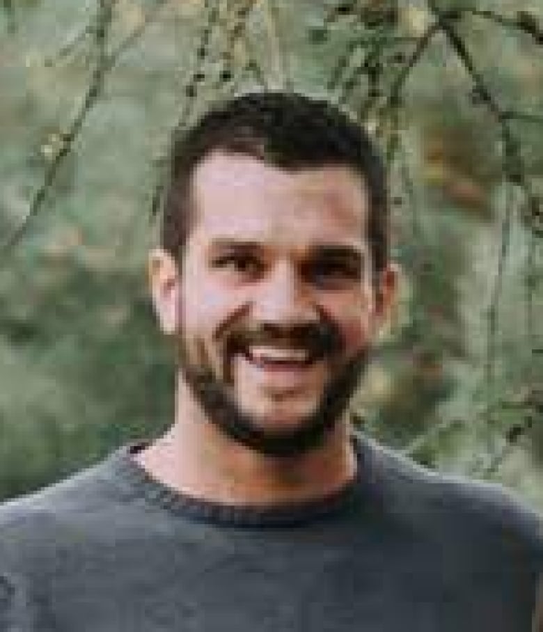 A man in about his 30s or 40s with short dark hair and a short beard and mustache smiles broadly at the camera, wearing a gray sweater. Branches hang against greenery in the background.