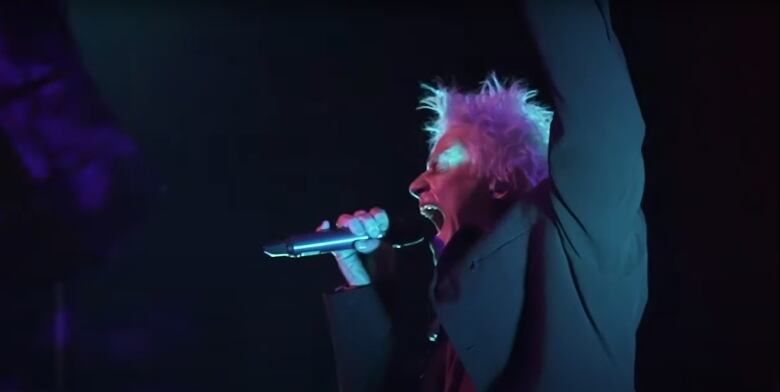 A man with spiky light-coloured hair sings into a microphone on a stage.