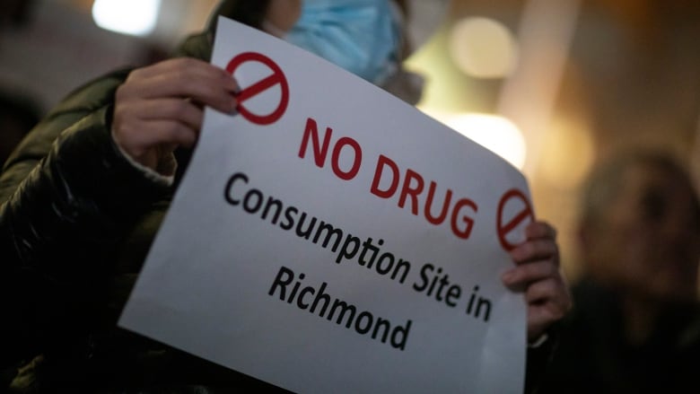 A woman wearing a facemask holds up a sign that reads 'No Drug Consumption Site in Richmond'.