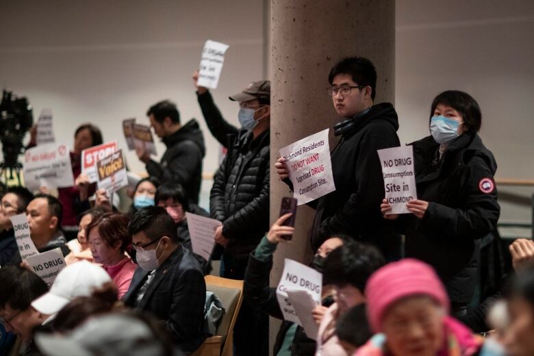 People hold up signs reading 'Richmond residents do not want supervised drug consumption site' and 'no drug consumption site in Richmond' in a city hall.