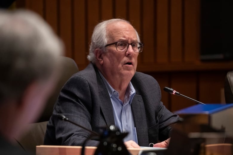 An old white man looks concerned as he sits in a city hall chamber.