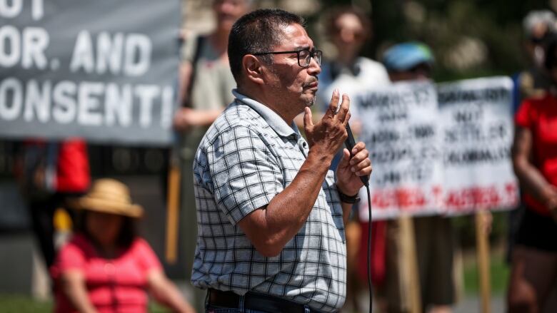 A man speaks into a microphone with demonstrators holding signs behind him.