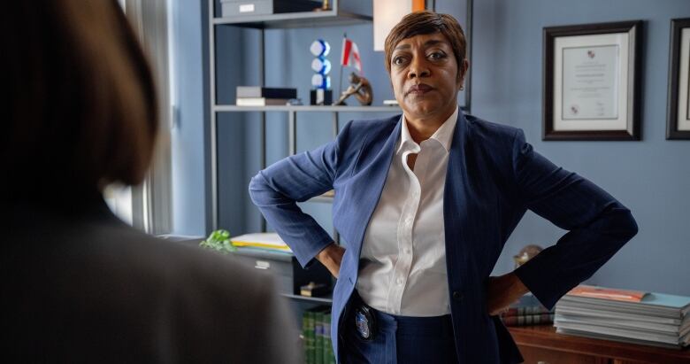 An image shows actress Karen Robinson on set of Law and Order in a blue suit, standing in an office with her hands on her hips. 