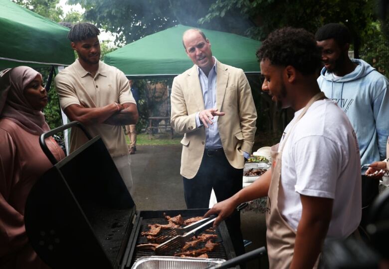 Several people stand around as someone cooks on a barbecue.