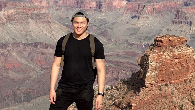 A young man smiles, standing against a scenic, canyon backrgound. 