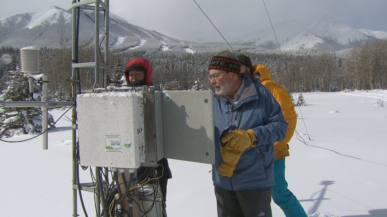 A man wearing glasses and a blue jacket inspects a box against a backdrop of the Rocky Mountains.