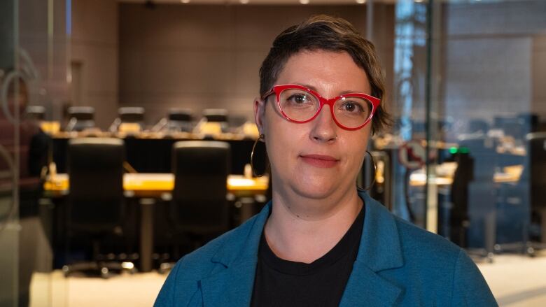 A woman with red glasses and a blue blazer stands in front of an empty room with many chairs.