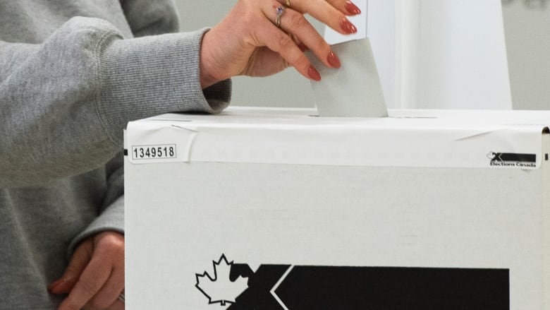 A hand holding a pencil puts a ballot into a voting box.