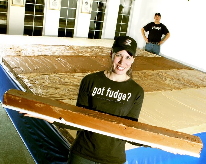 A woman wearing a ball cap holds a long piece of chocolate fudge in front of a large chocolate fudge bar on the ground behind her.