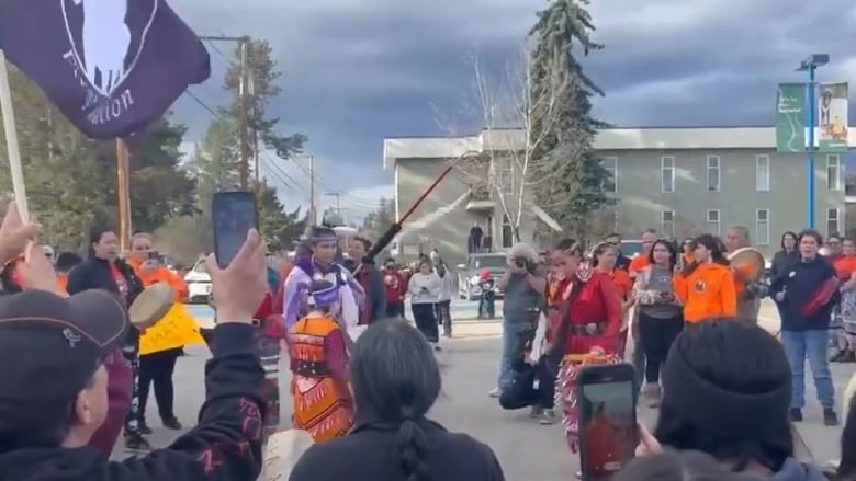 A crowd of people, some holding cell phones, flags, or drums, form a circle around Indigenous dancers wearing regalia, during a protest march to city hall.  