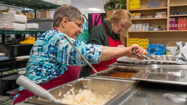 Two people scoop food out of metal trays at a serving station.