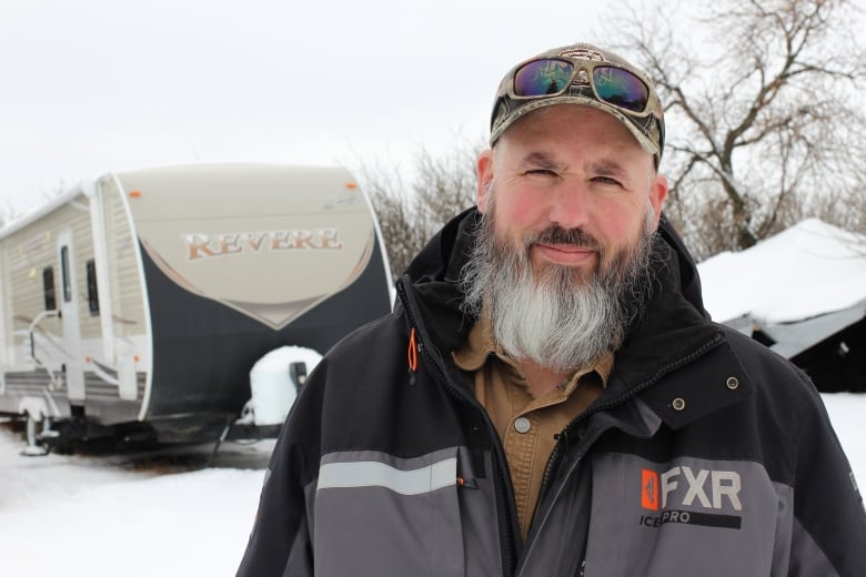 A man with a ball cap, sunglasses and a bear stands in front of an RV on a snowy day in Alberta.