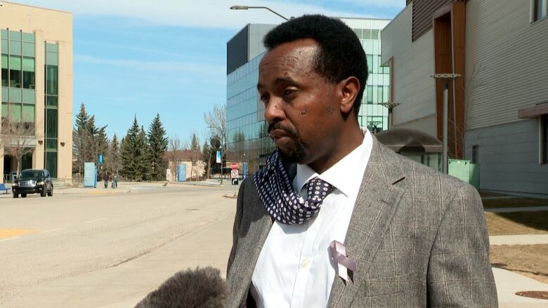 A man in a tie stands outside on a sidewalk, speaking into a microphone. 