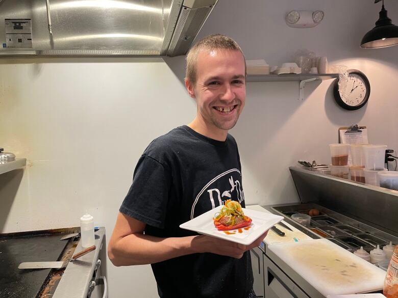 Smiling man in a black shirt holding a white plate with a stuffed tomato on it in an industrial kitchen