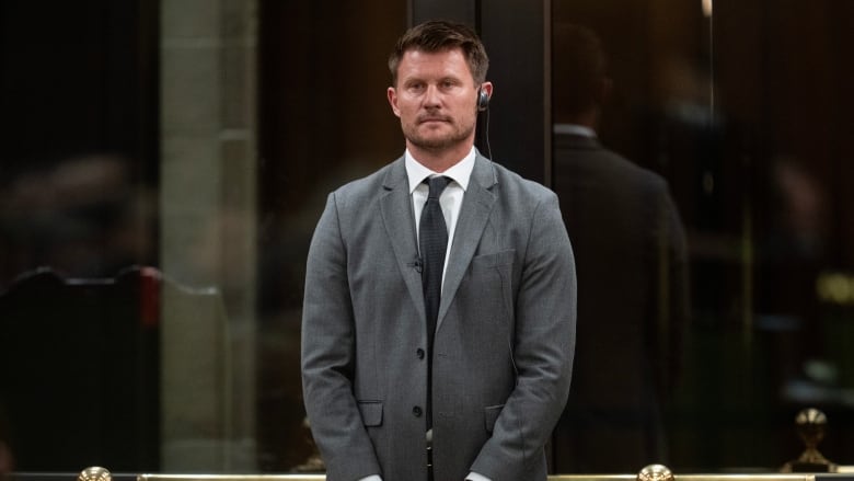 A man in a grey suit stands in front of the bar of the House of Commons.