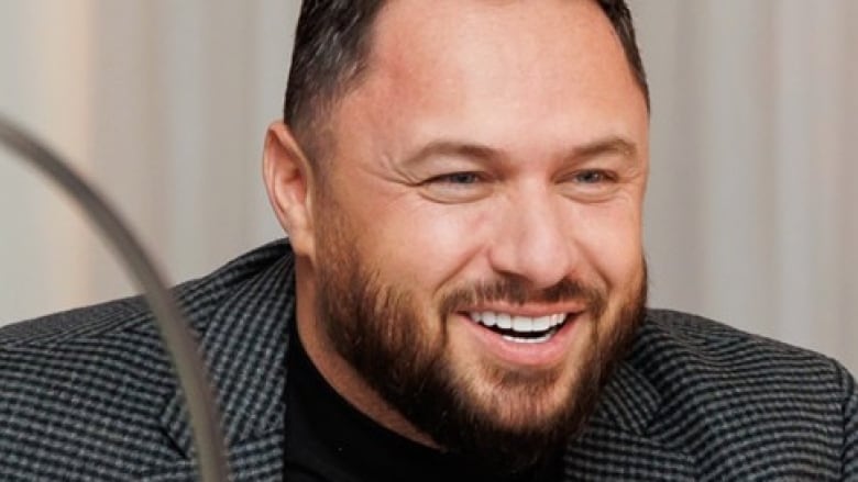 A smiling man with dark hair and a beard is seen wearing a dark checkered blazer. 