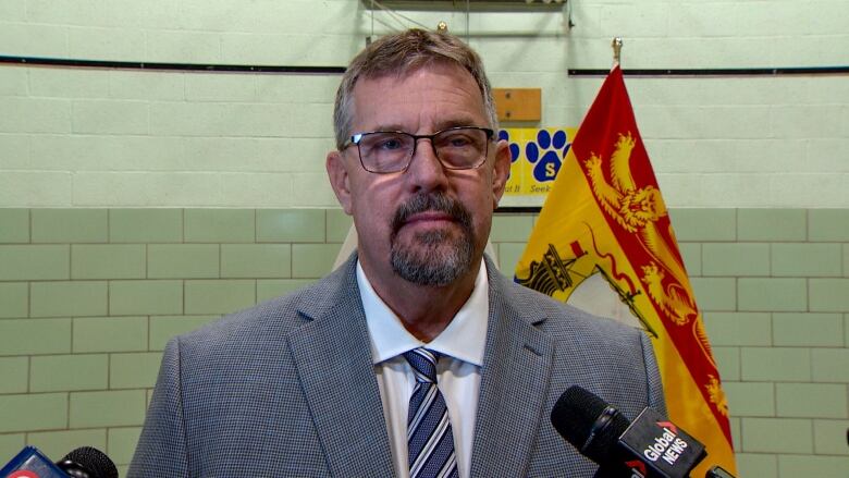 Man in suit and tie in front of a flag with microphones in front of him. 