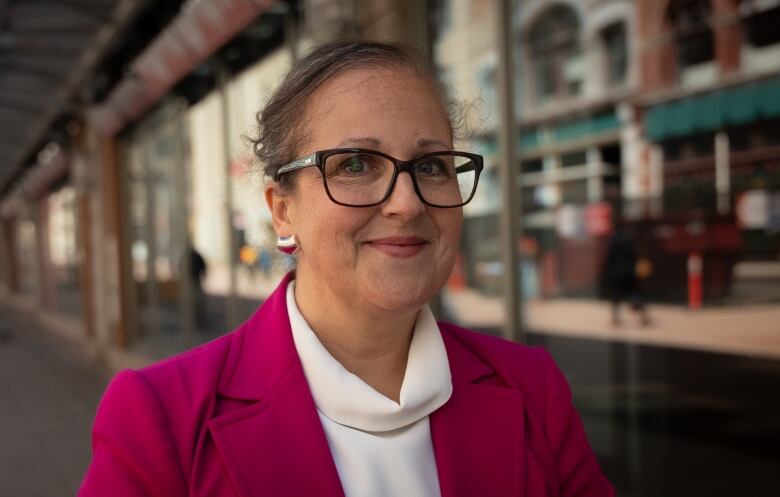 Carolyn Holland, a cancer survivor, smiles at a camera. She's wearing a bright pink blazer and glasses.