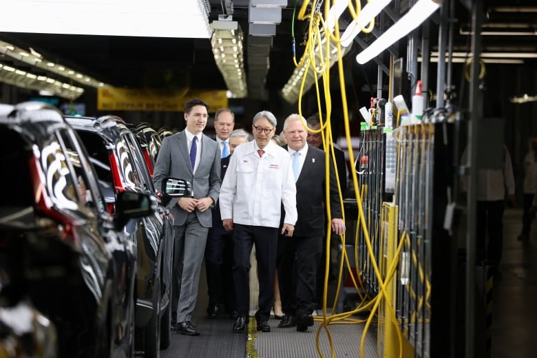 Three men walking inside a car assembly plant, with vehicles on one side and equipment on the other.