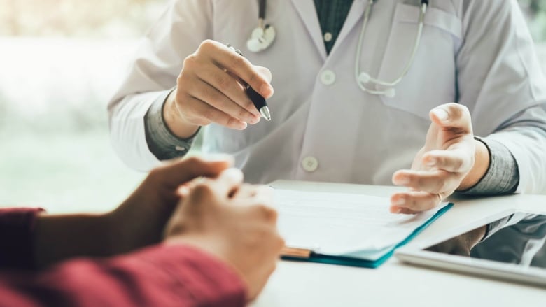 A stock photo showing a doctor's hands holding a pen over documents and a patient with his arms on the table sitting across from him. 