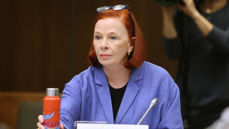 A woman in a lavender blazer sets a water bottle down onto a table.