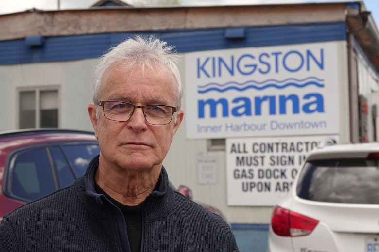 An older man with white hair and wearing glasses stands in front of of some parked cars and a sign that says 