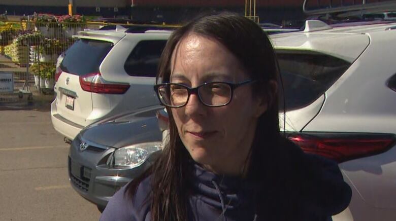 A white woman, with straight black hair, is wearing glasses and navy blue hoodie. She is standing beside her vehicle in a parking lot, under the sunshine.