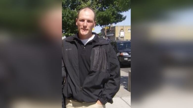 A man in a black jacket and light pants stands on the sidewalk.