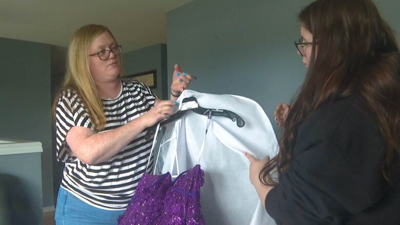 A woman with long blonde hair and a black and white striped shirt holds up a dress bag with the top of a purple dress showing. A teenage girl wearing a black hoodie appears on the left, also holding the bag.