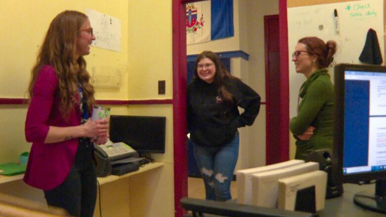 A teenage girl wearing a dark hoodie stands in the centre, chatting with a woman wearing a red blazer, left, and a woman with a green dress, right. 