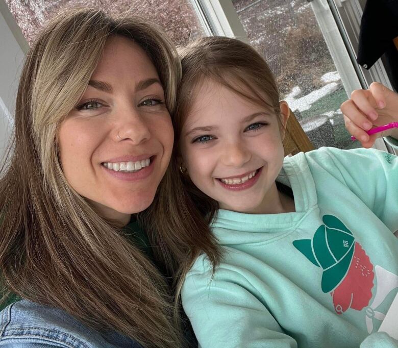 A woman with blonde hair smiles and poses with her eight-year-old daughter. Her daughter wears a light blue hoodie. 