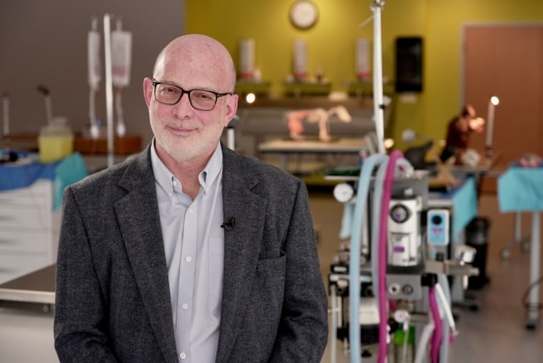 A bald man with white stubble, glasses, a white shirt and dark grey blazer stands in front of medical equipment and intravenous bags for animals.    