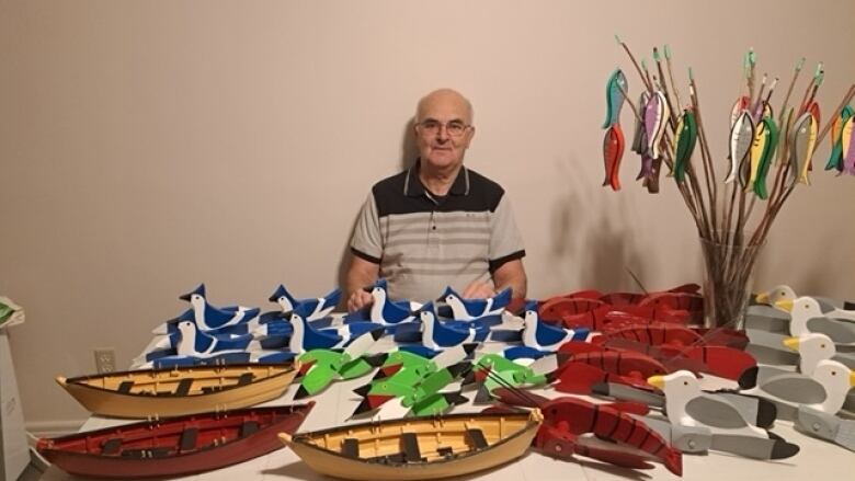 A man sits behind a table filled with wooden boats, seagulls, lobsters, blue jays, and and a handful of wooden fish hanging out of a vase.