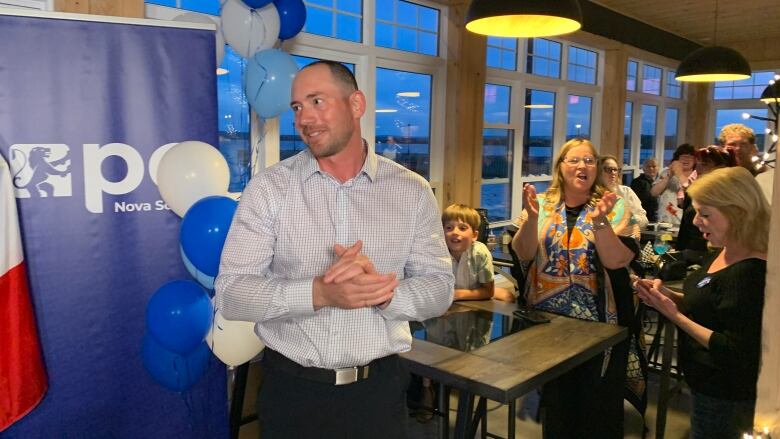 A man stands in front of a blue sign with blue balloons and people clapping nearby.