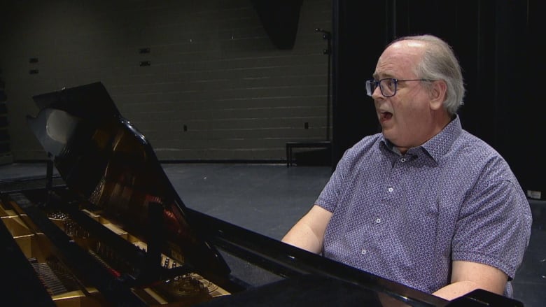 A man wearing a buttoned-up shirt with glasses sits at a piano.