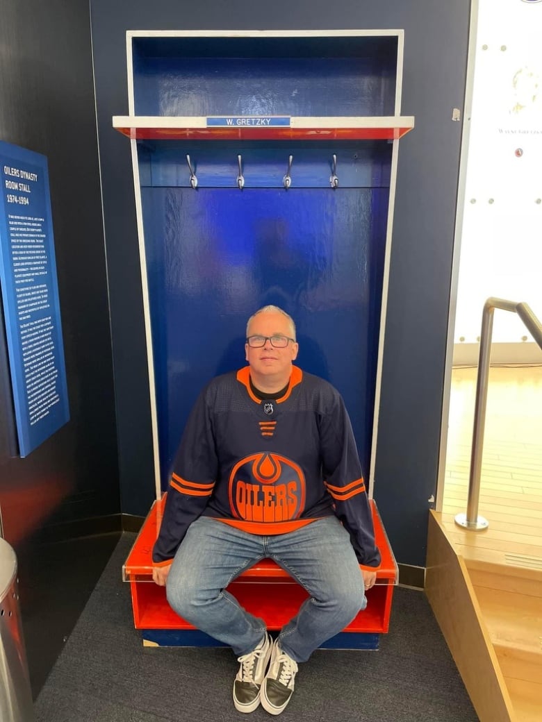 A man wearing an Edmonton Oilers jersey sits in Wayne Gretzky's former locker room stall now on display at Rogers Place.