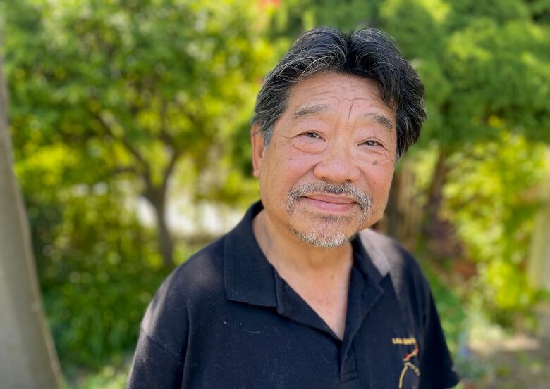 A man with black hair and a greying goatee in a black polo shirt stands in front of leafy trees. 