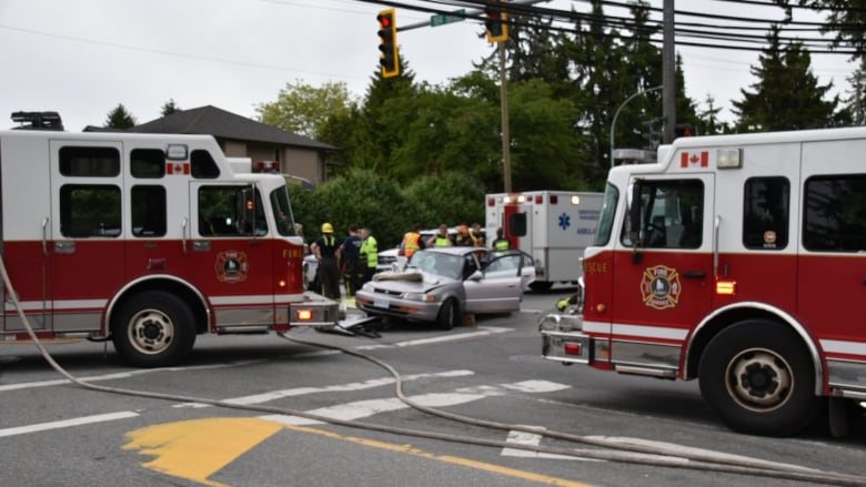 An ambulance, fire trucks, and paramedics are gathered in an intersection, where a damaged grey small car sits. 