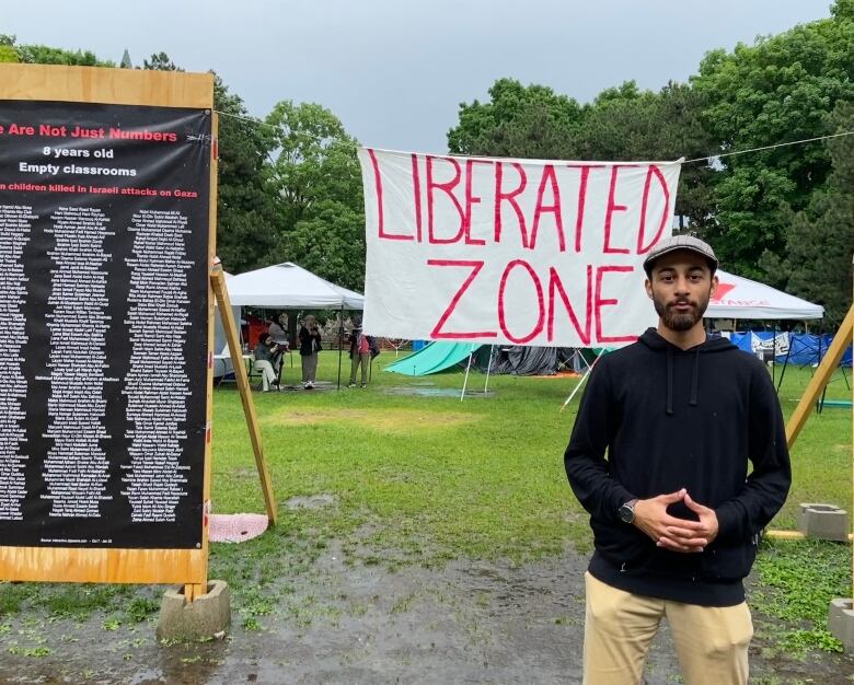 A man in a black hoodie and a grey cap stands in front of a sign at the University of Ottawa's Tabaret Lawn that reads 