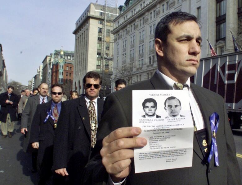 A man in a suit is shown in the foreground leading others who are similarly dressed, walking down a sidewalk. He holds a picture of two men.