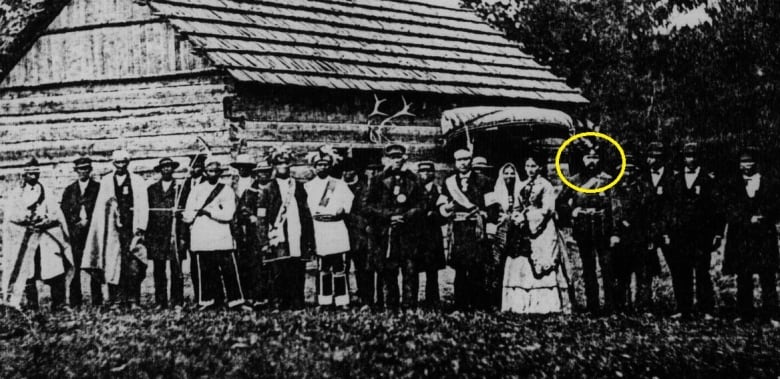 A row of Six Nations men in ceremonial regalia stand outside a wooden structure.