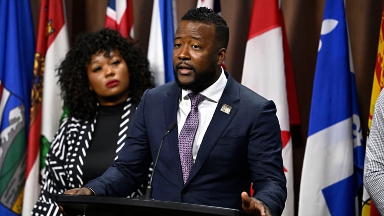 A man in a suit speaks at a podium into a microphone while a woman stands beside him.