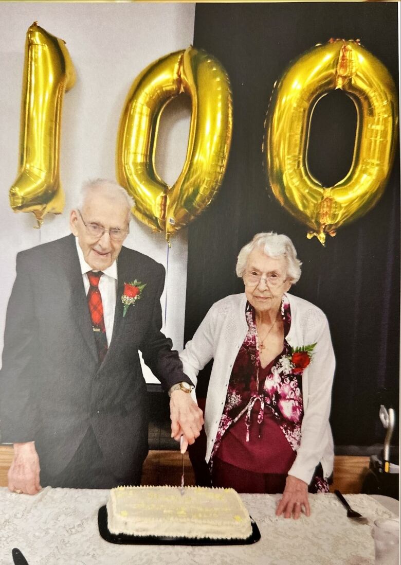 A man and women hold a knife, about to cut birthday cake, with balloons spelling the number 100 behind them.