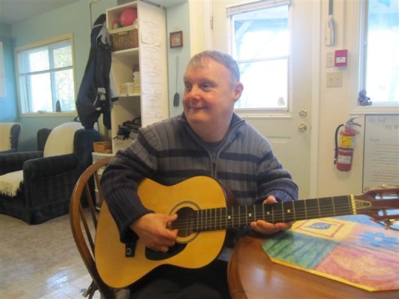 A man sits in a chair strumming a guitar at a kitchen table. 