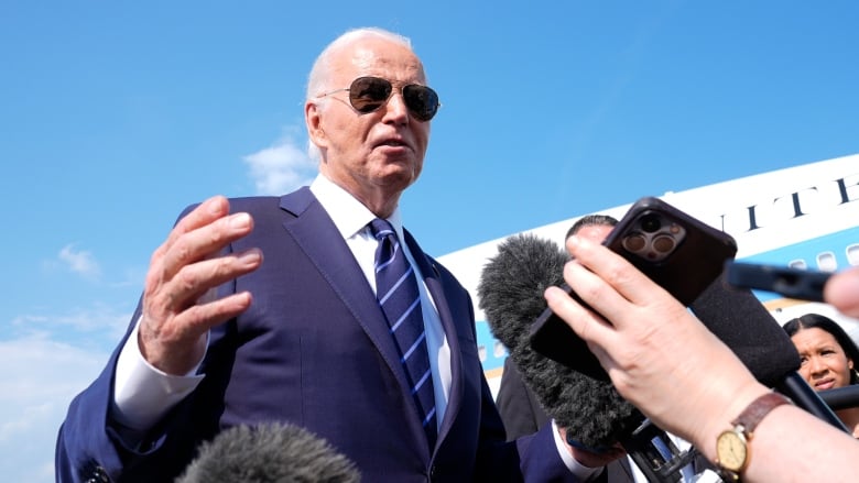 President Joe Biden, wearing dark sunglasses, a blue suit and tie, stands before a crowd reporters holding microphones in front of a large, white passenger jet. 